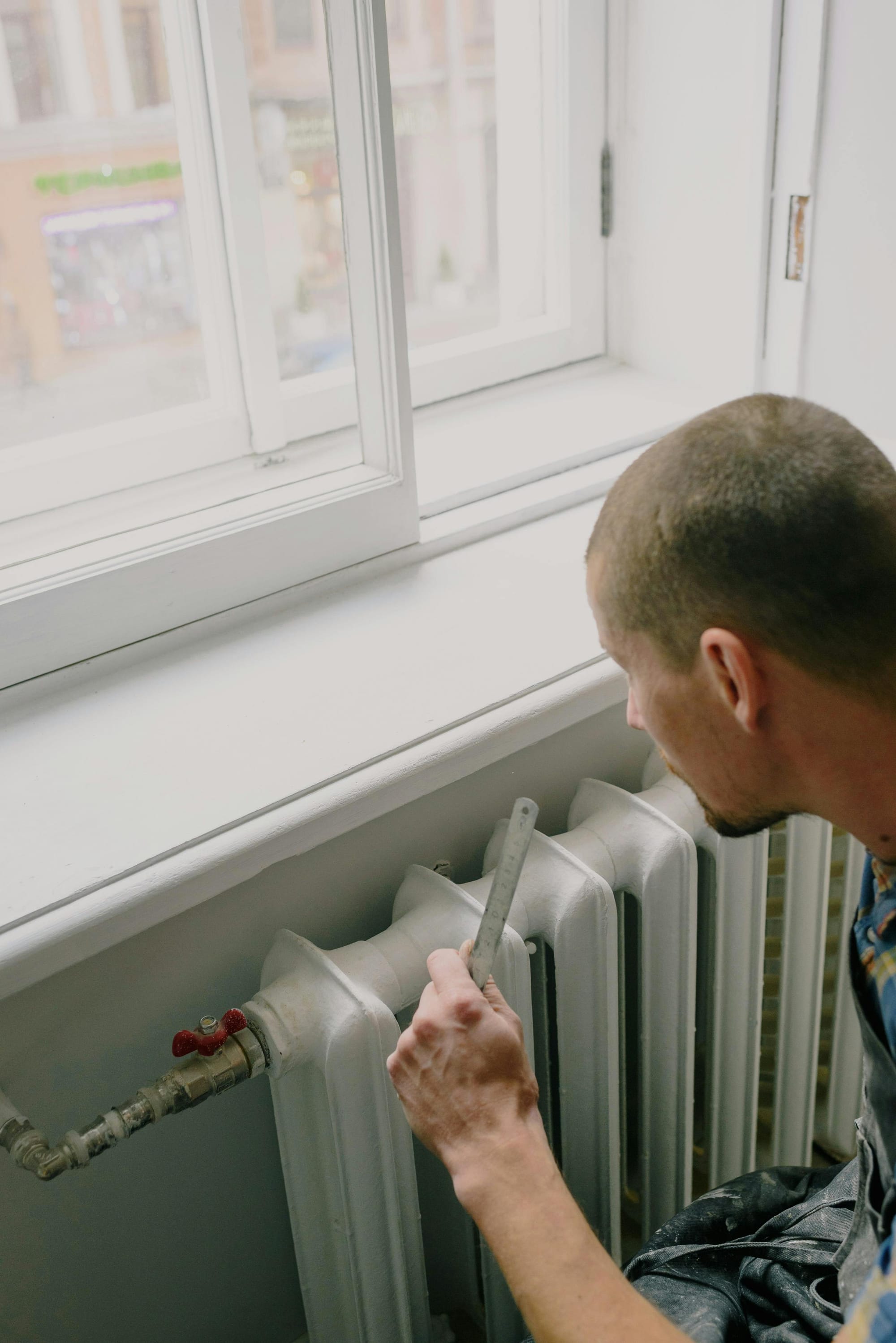Technician bleeding a cast-iron radiator beneath a window during home heating maintenance