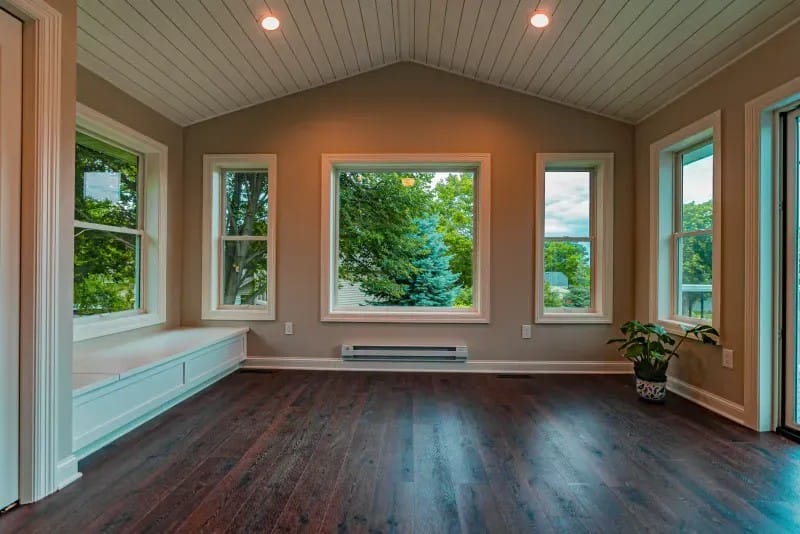 Bright sunroom with vaulted ceiling, large windows, built-in bench, and view of lush greenery.