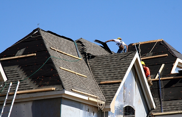 Construction workers installing asphalt shingles on a multi-gable residential roof under clear blue sky.
