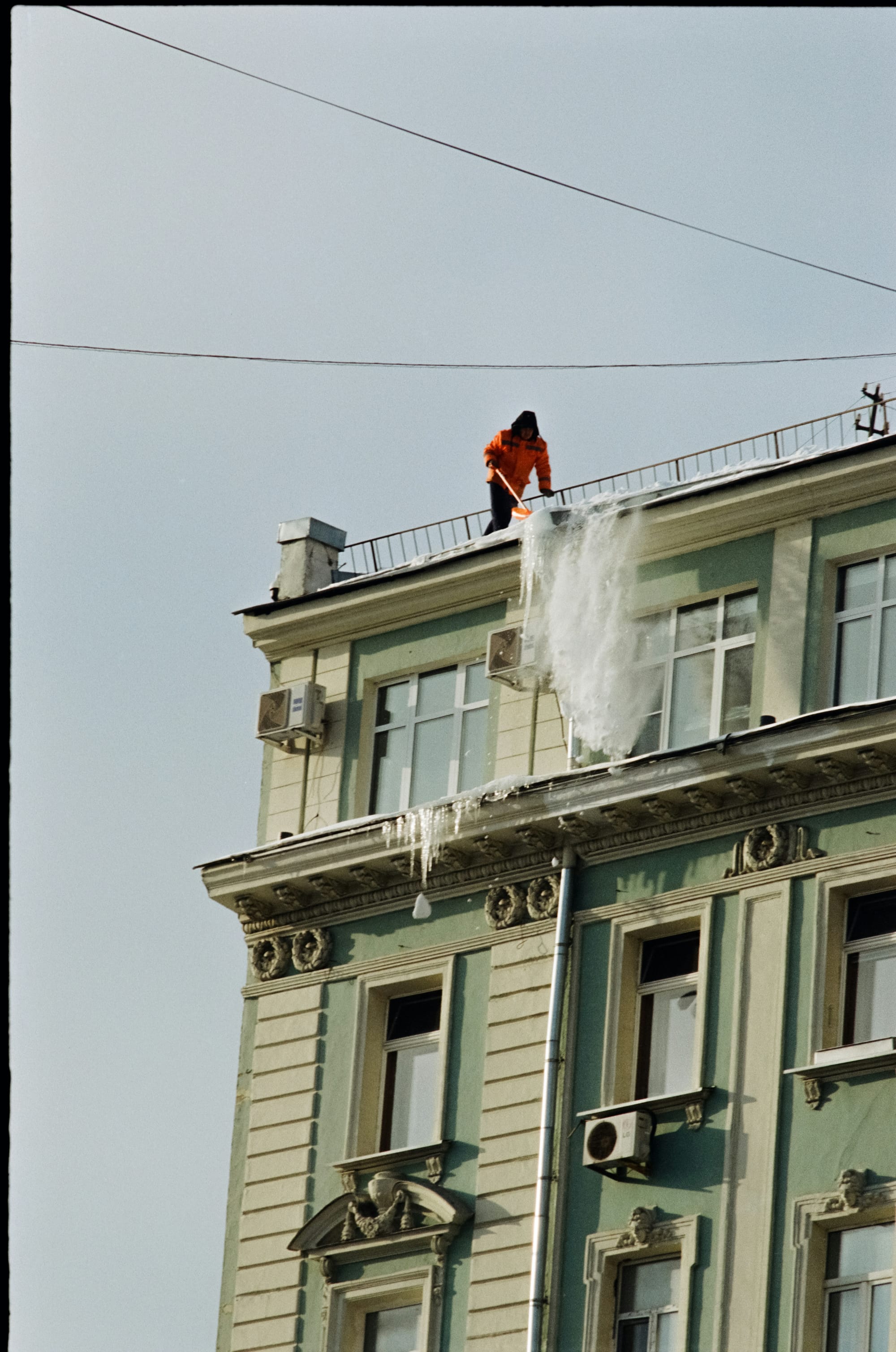 Person in orange jacket clearing heavy icicles from ornate building roof during winter maintenance.