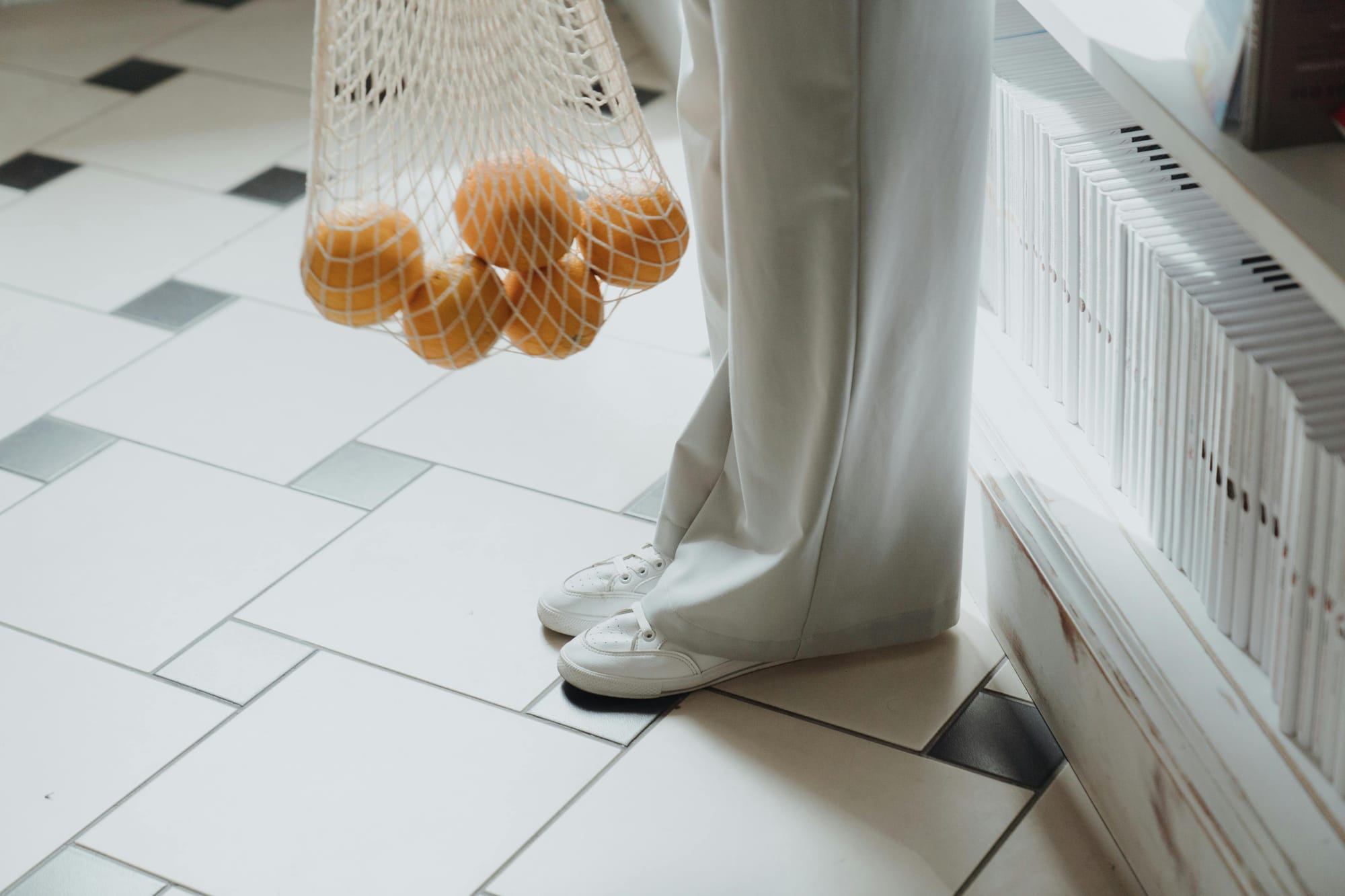 Person holding a mesh bag of oranges, standing on tiled floor near shelf of white books.