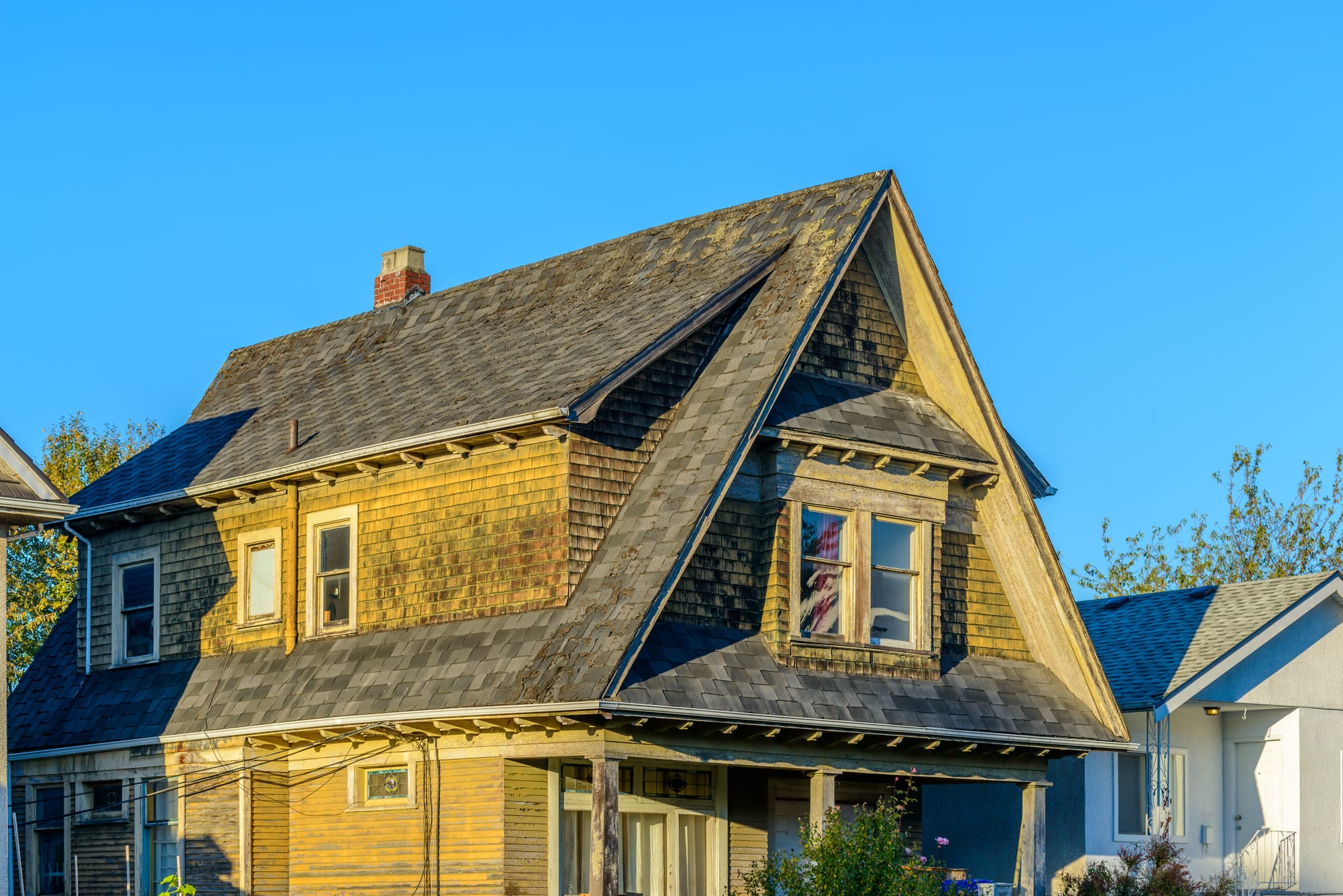 A weathered two-story house with wood siding, steep roof, and covered porch, showing signs of age beside a modern building.