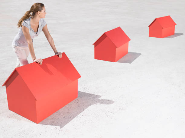 Person leaning on a large red house model with smaller houses trailing behind, symbolizing housing scale or market progression.