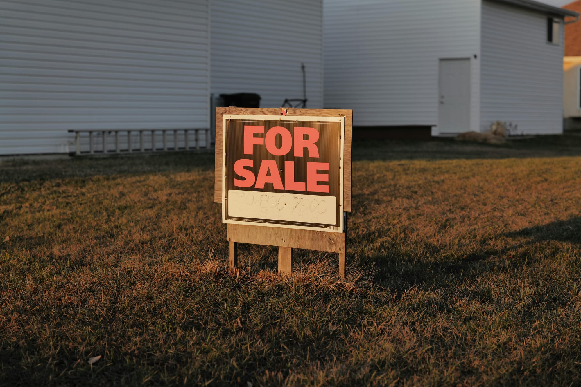 For Sale sign on a grassy lawn in front of a white house with siding and a ladder leaning against the wall, photographed in warm evening light.