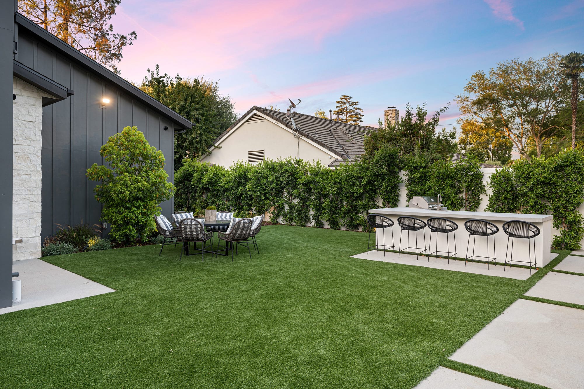 Modern backyard with artificial grass lawn, outdoor dining table, and sleek kitchen bar with grill and bar stools, surrounded by tall hedges at sunset.