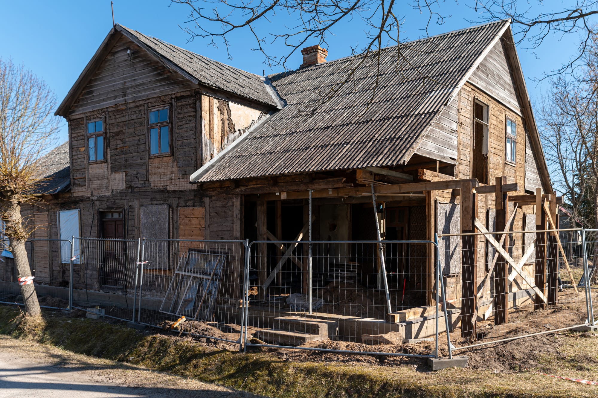 Old wooden house under renovation with boarded windows, corrugated roof, and temporary beams supporting the porch, surrounded by construction materials and fencing.