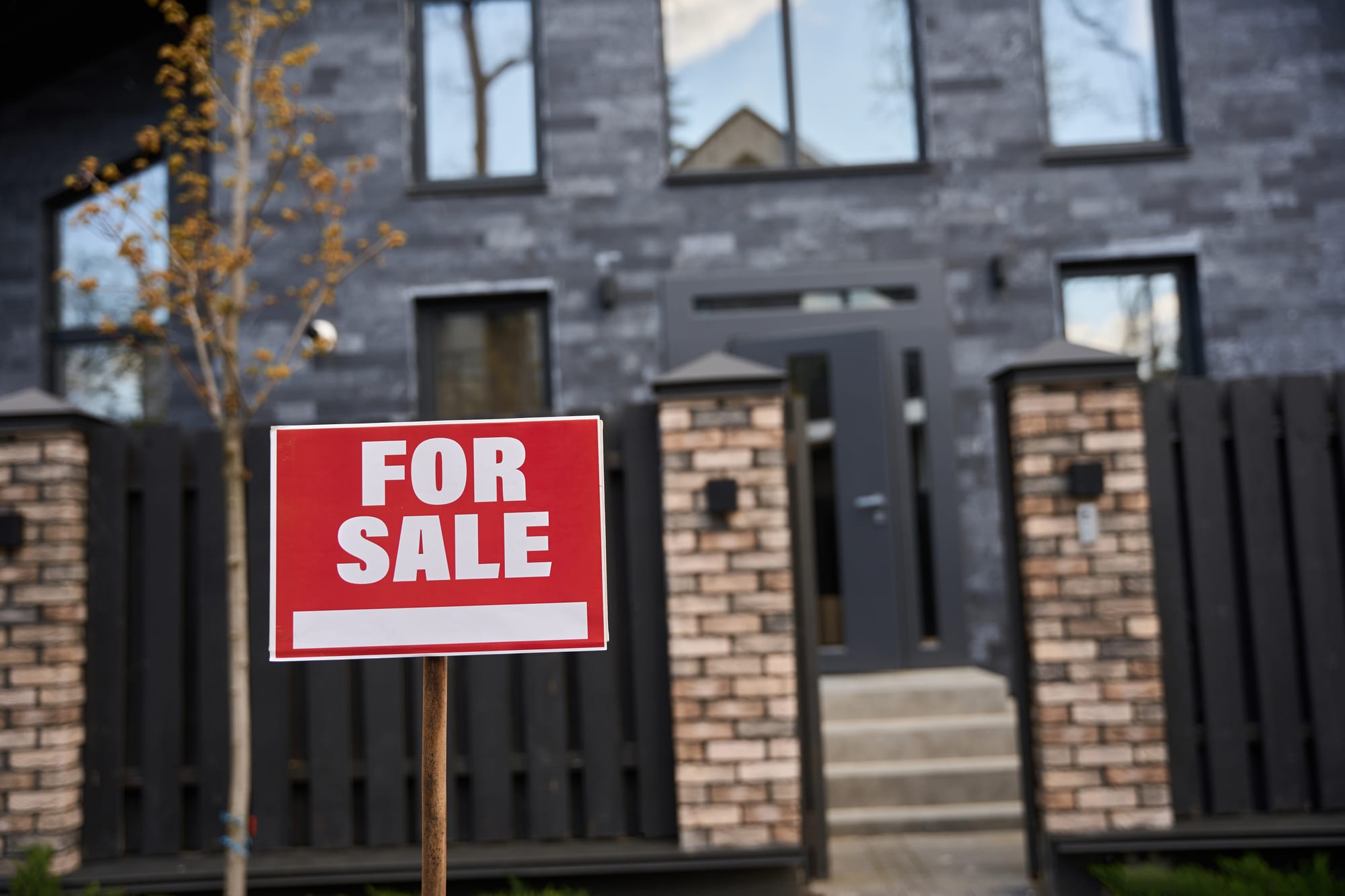 A red "FOR SALE" sign standing in front of a modern, dark-tiled house with a brick and wood fence.
