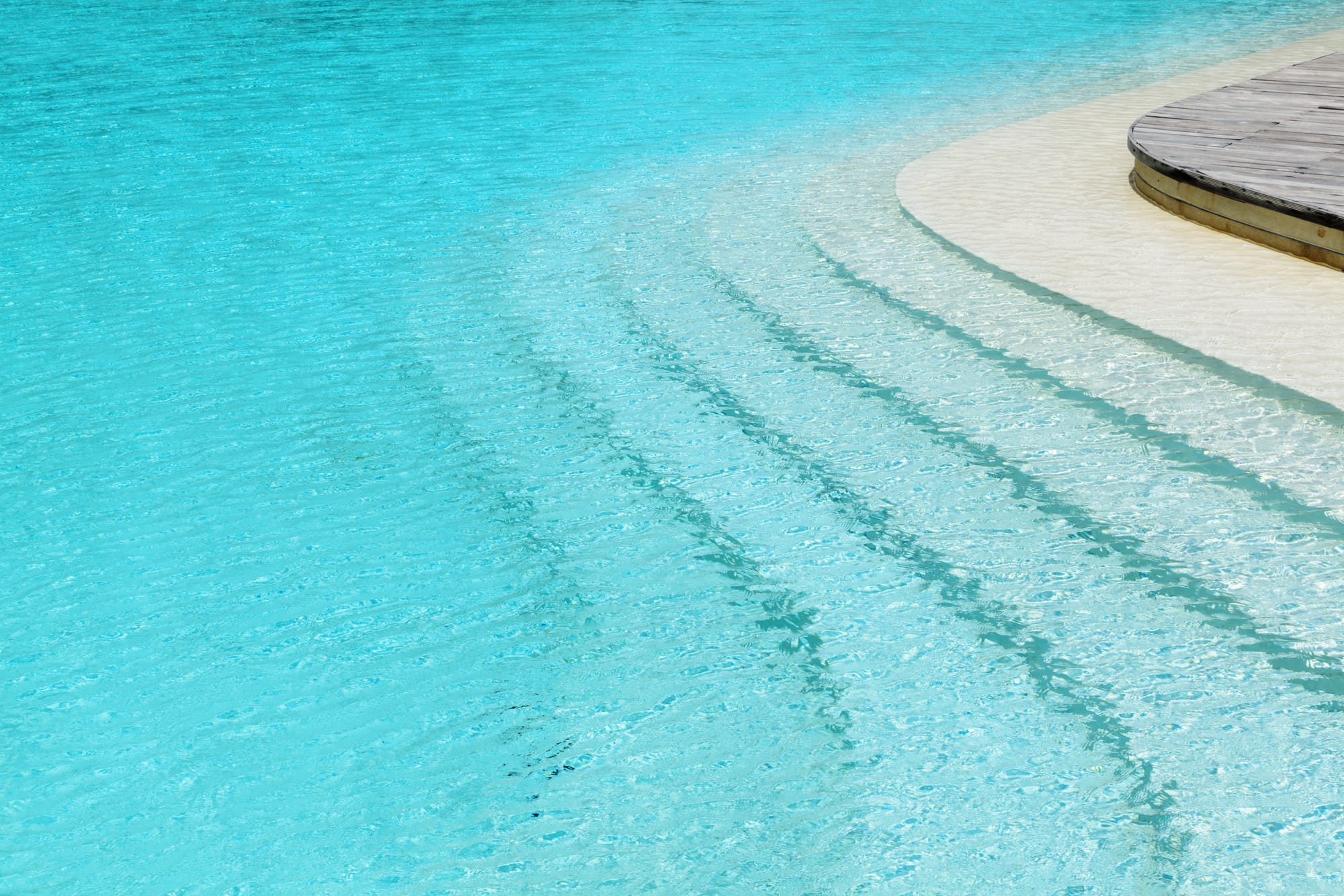 A high-angle view of wide, curved steps descending into a turquoise swimming pool with clear, rippling water. A section of a dark wood deck is visible in the upper right corner.
