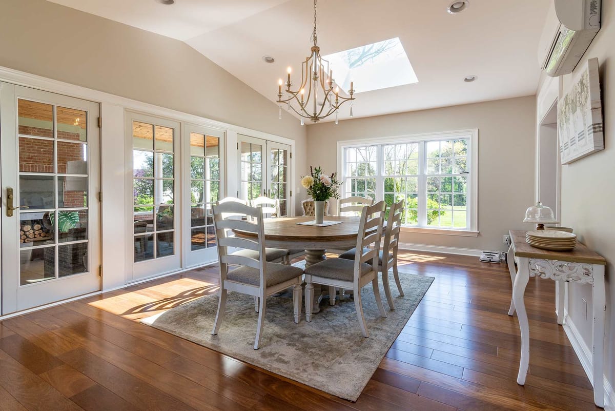 Elegant dining room with round table, skylight, chandelier, and large windows overlooking greenery.
