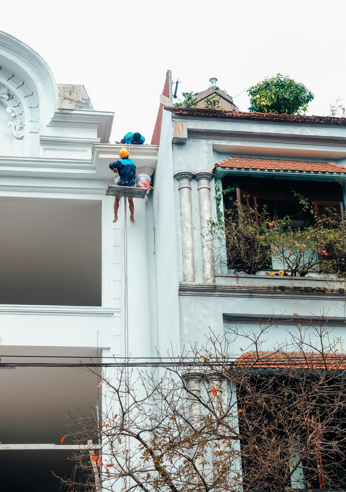 **Alt text:** Worker in a yellow hard hat hangs from a rope platform, painting the upper facade of a white building beside a blue house with a plant-filled balcony.