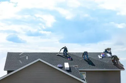 Four workers installing asphalt shingles on a gable roof under a partly cloudy sky.