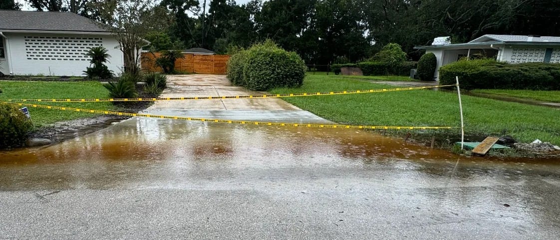 Flooded residential driveway cordoned off with yellow caution tape; shallow water pooling across entrance and street.