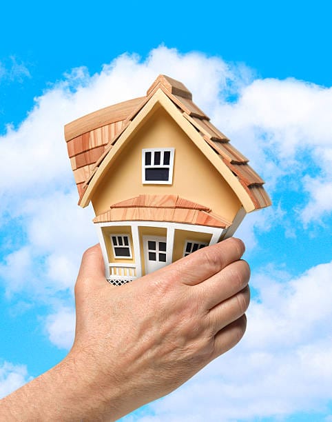 Hand squeezing a small yellow house model against a blue sky, symbolizing housing stress or market pressure.