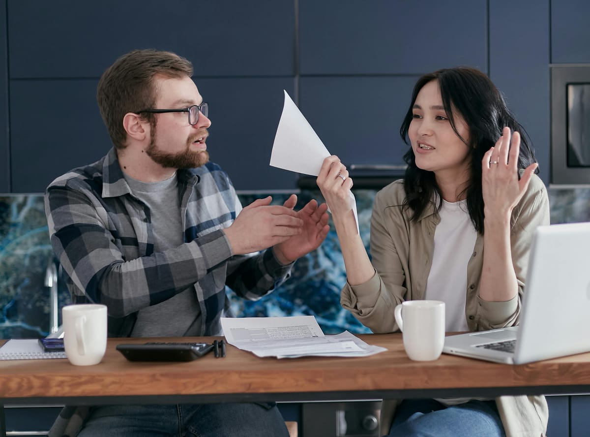 Two homeowners seated at a kitchen table reviewing paperwork and a laptop, looking focused and tired