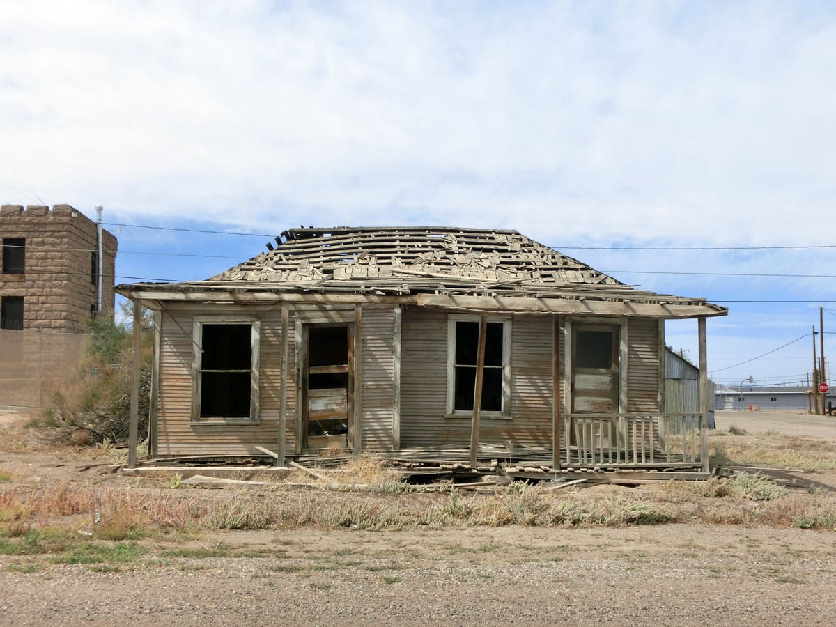 Old abandoned wooden house with collapsed roof, broken porch, and missing windows, set in a rural town with utility poles and sparse vegetation.