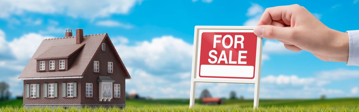 A miniature model house next to a hand holding a red "FOR SALE" sign on a grassy field.