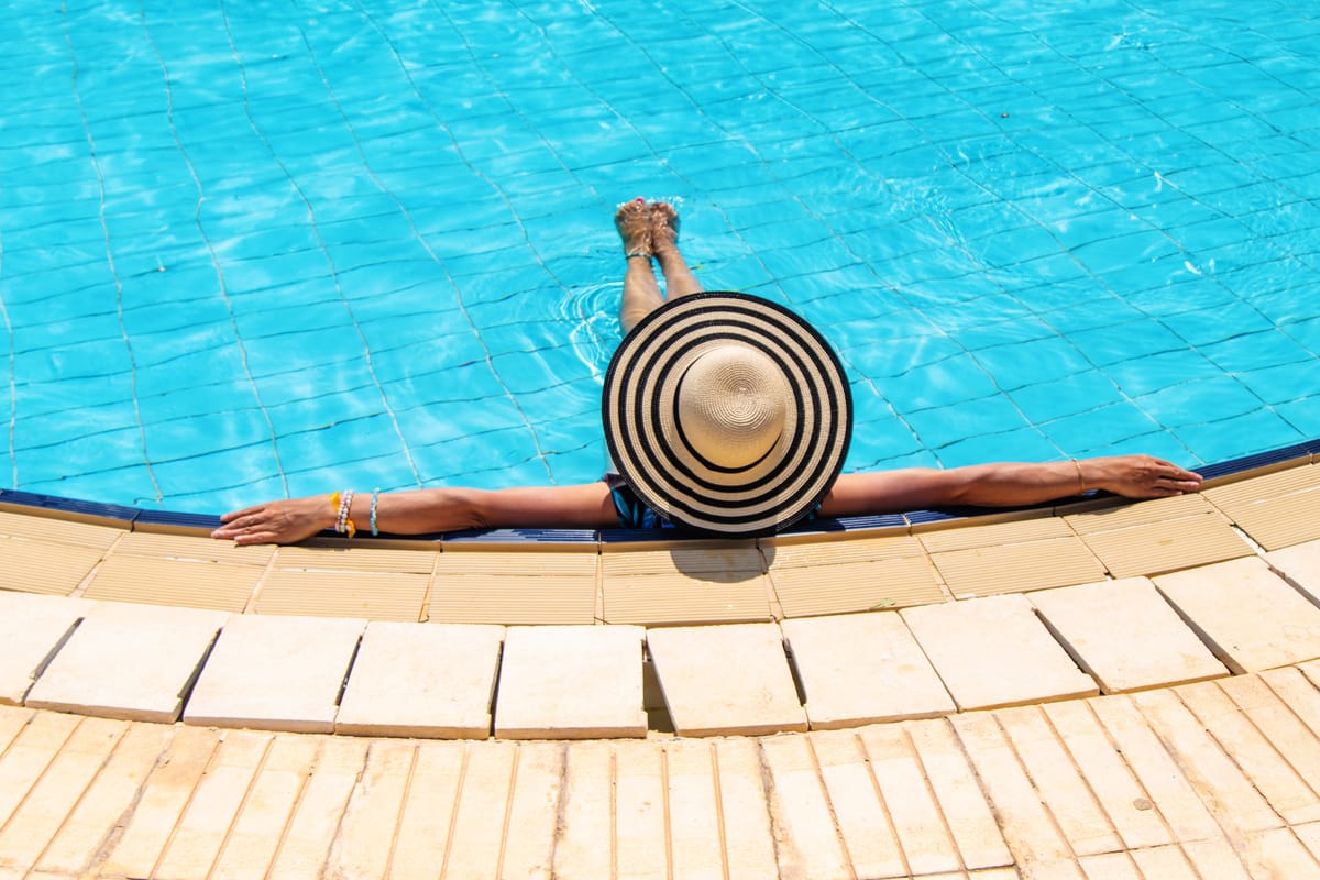 An overhead view of a person wearing a large black-and-white striped sun hat, relaxing at the edge of a bright blue tiled swimming pool with their arms outstretched along the stone coping.