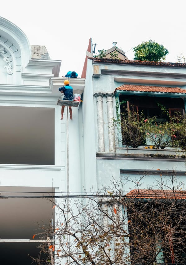 **Alt text:** Worker in a yellow hard hat hangs from a rope platform, painting the upper facade of a white building beside a blue house with a plant-filled balcony.