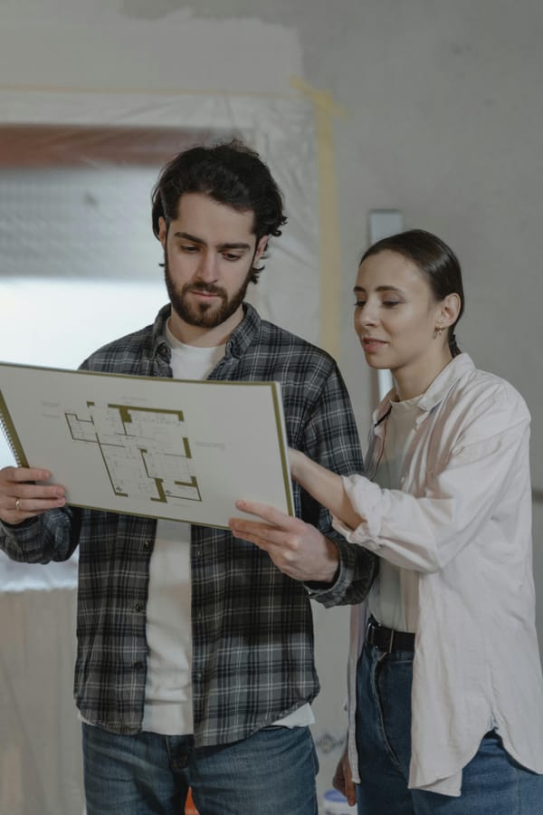 “Two homeowners stand indoors reviewing a floor plan as they plan a home remodel.”