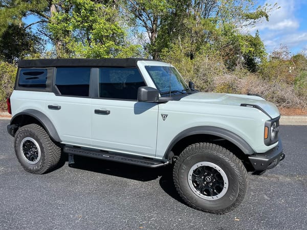 Light gray Ford Bronco with black soft top and off-road tires parked near trees.