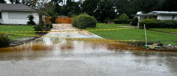 Flooded residential driveway cordoned off with yellow caution tape; shallow water pooling across entrance and street.