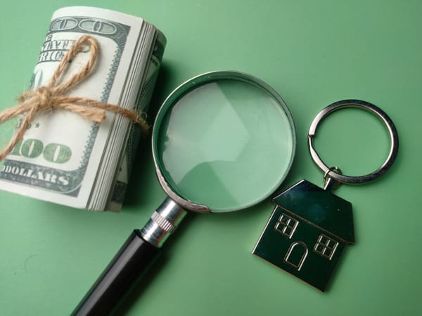 Bundle of $100 bills with house keychain and magnifying glass on green background.