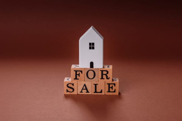 Small white model house on wooden blocks spelling FOR SALE against warm brown background.