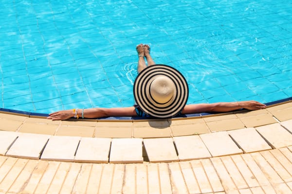 An overhead view of a person wearing a large black-and-white striped sun hat, relaxing at the edge of a bright blue tiled swimming pool with their arms outstretched along the stone coping.